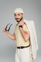 Fashionable gay man in hat holding sunglasses and jacket isolated on grey.