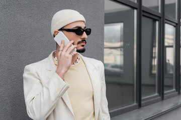 Trendy homosexual man in sunglasses talking on mobile phone on urban street.