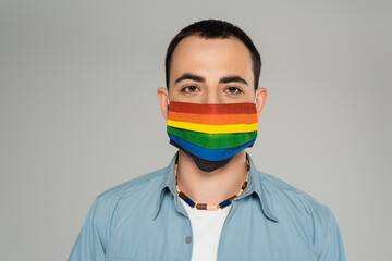 Portrait of brunette homosexual man in medical mask with lgbt flag isolated on grey.