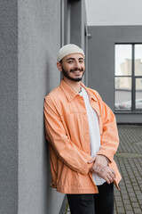 Stylish gay man in hat and jacket smiling at camera near building on urban street.