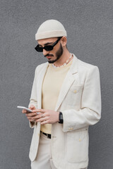 Trendy gay man in sunglasses using smartphone near wall outdoors.