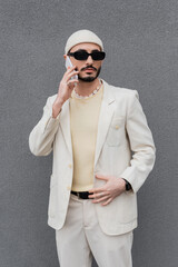 Fashionable gay man in suit and sunglasses talking on smartphone near building outdoors.