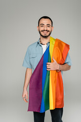 Young bearded gay man with lgbt flag smiling at camera isolated on grey, international homophobia...