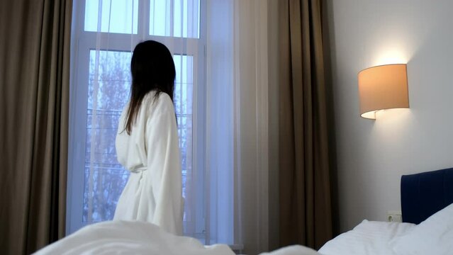 Brunette young slim woman in white bathrobe is looking at window on winter trees in hotel room. She is smiling and standing near the bed. Vaction, relax and rest with comfort in winter concept.