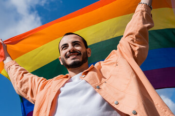 Low angle view of cheerful gay man holding lgbt flag with sky at background outdoors, International...