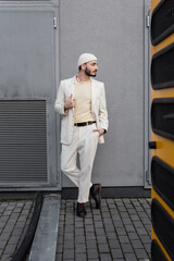 Stylish gay man in hat and suit posing near building on urban street.