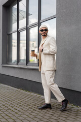 Gay man in stylish suit and sunglasses holding coffee to go on urban street.