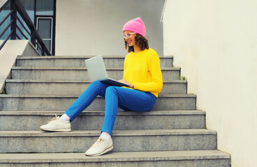 Fototapeta na wymiar Portrait of stylish modern young woman working with laptop wearing a colorful clothes, eyeglasses in the city