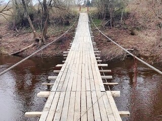 Fototapeta premium Suspended wooden pedestrian bridge to the other side of the river in the forest. View of the suspension bridge in the forest. 