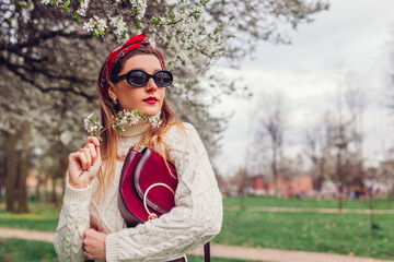 Portrait of woman wearing red hair scarf and sunglasses in spring park. Retro female fashion. Headscarf and purse