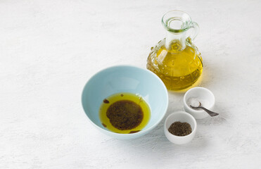 A blue bowl with olive oil, spices and balsamic sauce surrounded by a bottle of olive oil, a salt and pepper shaker on a light gray background. Traditional salad dressing preparation