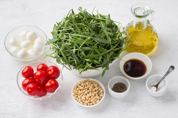 Ingredients for a delicious salad: arugula, cherry tomatoes, mini mozzarella, pine nuts, olive oil, balsamic sauce, salt, black pepper on a light gray background