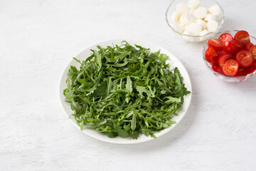 Plate with fresh arugula leaves, bowls with chopped cherry tomatoes and mini mozzarella on a light gray background. Cooking delicious vegetarian salad