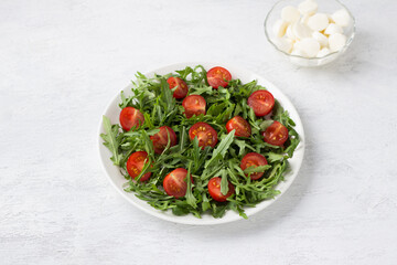 Plate with fresh arugula leaves, chopped cherry tomatoes and bowl mini mozzarella on a light gray background. Cooking delicious vegetarian salad