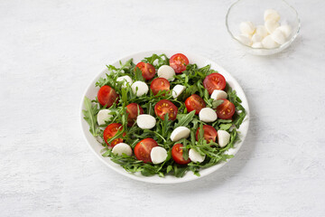 Plate with fresh arugula leaves, chopped cherry tomatoes and mini mozzarella on a light gray background. Cooking delicious vegetarian salad