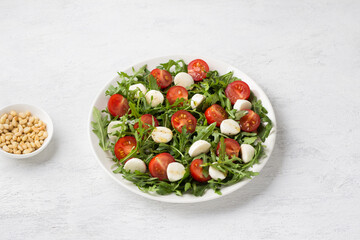 Plate with fresh arugula leaves, chopped cherry tomatoes and mini mozzarella sprinkled with salad dressing and a bowl of pine nuts on a light gray background. Cooking delicious vegetarian salad