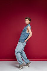full length of stylish model in denim outfit with vest posing with hands in back pockets on jeans on burgundy background.