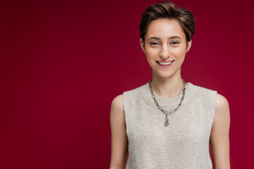 pleased young woman in sleeveless shirt and chain necklace standing on maroon background.