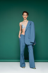 full length of young model with short hair posing in satin bra with blue blazer on shoulder on turquoise green background.