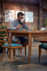 Young hipster man with earphones, siting in cafeteria, drinking coffee. Scrolling through his phone.