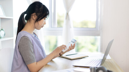 Asian woman working at home, Press the calculator to calculate the income - expenses of the house and use the laptop to record the information and check the accuracy, Home lifestyle.