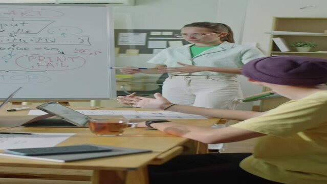 Vertical shot of gen Z girl pointing at computer algorithm written on whiteboard and giving presentation to IT team during office meeting