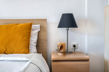 Standard bedroom details. Part of bed with dark yellow pillow on it and bedside table with standing lamp and candle. White background. 