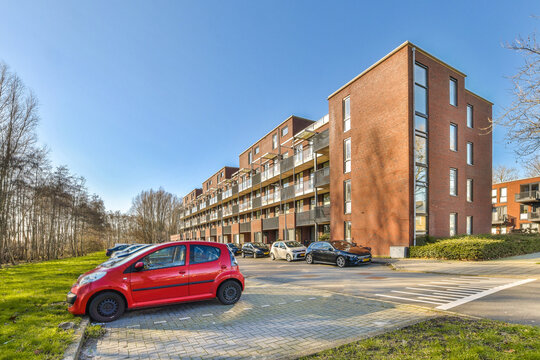 a red car parked in front of a large brick apartment building on a sunny day with blue sky and white clouds