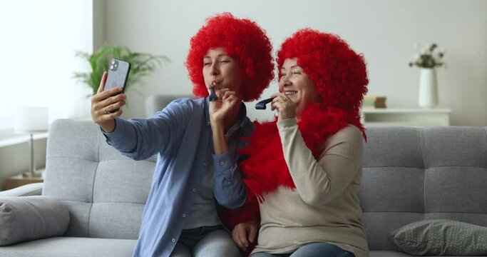 Happy adult daughter and older mother having fun, sit on sofa wear boa and red funny wigs blowing party whistles making selfie staring at smartphone camera, enjoy festive mood, celebrate holiday event