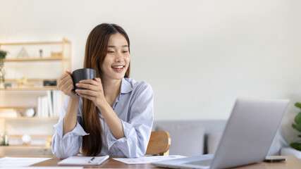 Beautiful Asian woman working on laptop and sipping coffee with smiling face in her home, Creating happiness at work with a smile, Freelancer working at home happily, Work from home.