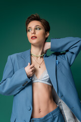 young woman with short hair posing in jacket with silk bra underneath on turquoise green.