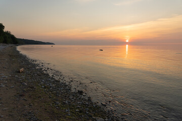 Fototapeta premium Summer sunset on the shore of the bay in Toila Oru Park. Orange clear sky. Ripples on the water from the wind. Pebbles on the shore. Natural background. Space for text.