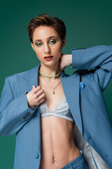 young model with short hair posing in blue jacket with silk bra underneath on turquoise background.