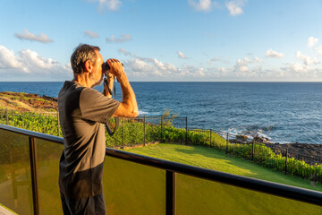 Mature Caucasian man using binoculars standing on a balcony over the ocean and green grass and iron...