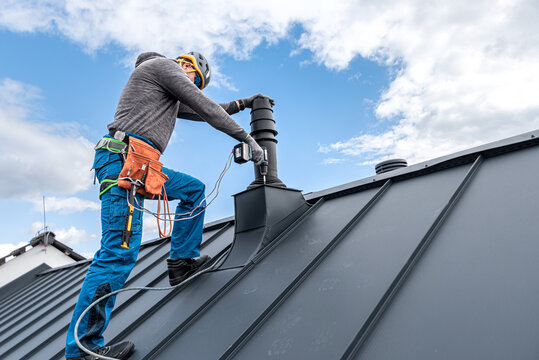 Work at heights. A tinman with tool belt and climbing harness on the roof is working with a cordless drill on the chimney sheeting.