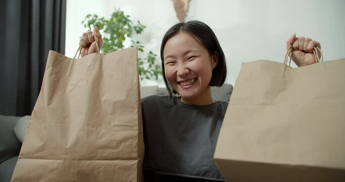 Excited asian lady shopping online, lifting gift bags above her head at home