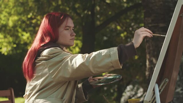 Focused Caucasian teenage girl with bright red hair painting on canvas outdoors in park lit by sunlight