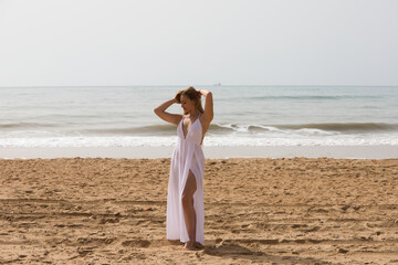 Fototapeta na wymiar Young beautiful blonde woman in white dress is walking on the sand on the shore of the beach on a sunny day. The woman makes different body expressions. In the background the blue sea.