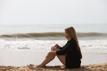 Fototapeta na wymiar Beautiful young blonde woman in black shirt sits on the shore of the beach sad and depressed. The woman is pensive and looks at the ground exhausted and tired. The woman is suffering.