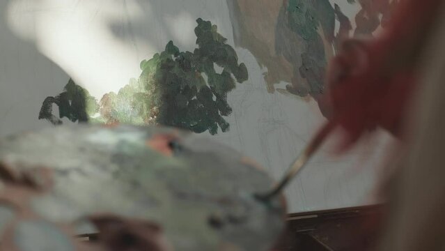 Closeup of unrecognizable female artist painting green landscape on canvas lit by sun rays outdoors