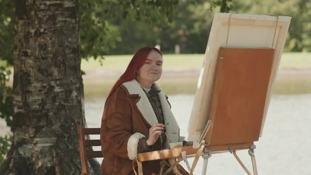 Medium portrait of Caucasian teenage girl with bright red hair smiling at camera while painting on canvas in summer park