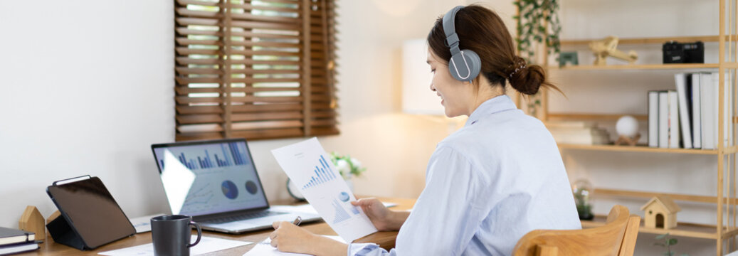 Asian female college student wearing headphones watching live performance or video call teacher teaching on laptop, Conversations with teachers and classmates, Online learning, Study at your own home.