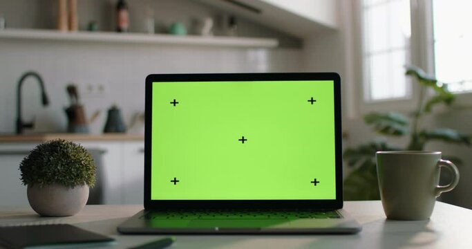 Laptop with green screen on display on kitchen table. Mockup of laptop with chromakey screen. Laptop on table is configured for work, business and study, mockup template for inserting videos or photos