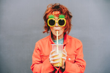 Hipster fashion young woman in bright clothes and pineapple glasses drinking fruity sugar flavored tapioca pearl bubble tea with straw on the gray wall background. Selective focus. Copy space. - obrazy, fototapety, plakaty