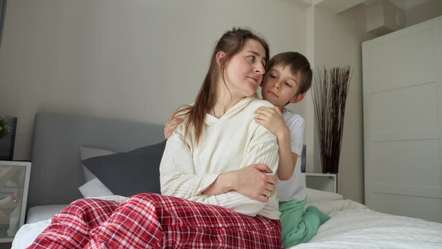 Little boy in pajamas hugging his sad mother on bed. Concept of loving family, parents and kids, love and support