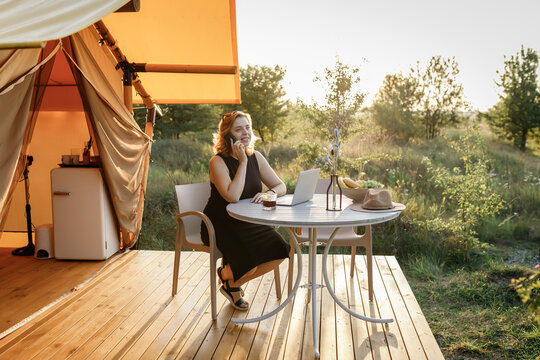 Smiling Woman freelancer talking by phone while working in cozy glamping tent in a sunny day. Luxury camping tent for outdoor summer holiday and vacation. Lifestyle concept