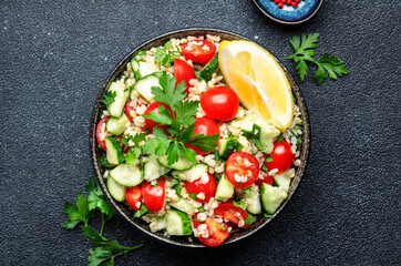 Bulgur tabbouleh salad with red cherry tomatoes, cucumbers, parsley and lemon dressing. Traditional Middle Eastern and Arabic dish. Bllack kitchen table background, top view