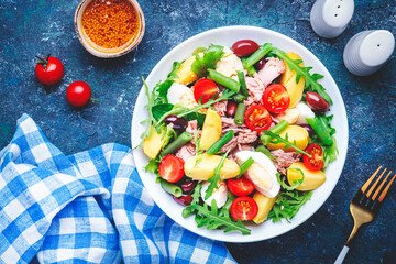 Nicoise salad with tuna, tomatoes, eggs, green beans, potatoes and olives on plate, blue table background, top view - obrazy, fototapety, plakaty