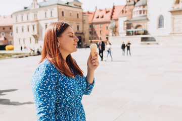 Fototapeta premium Happy young redhead woman with delicious white ice cream in waffle cone outdoors. Beautiful girl enjoying sweet food on historical building background, summer time