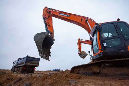 Ust-Luga, Leningrad oblast, Russia - November 16, 2021: Excavators Doosan and Hitachi loads dump truck. Rainy day, dirty ground on construction site.